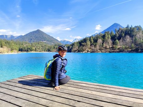 Person sitzt auf einem Holzsteg am blauen See, mit Bergen im Hintergrund.