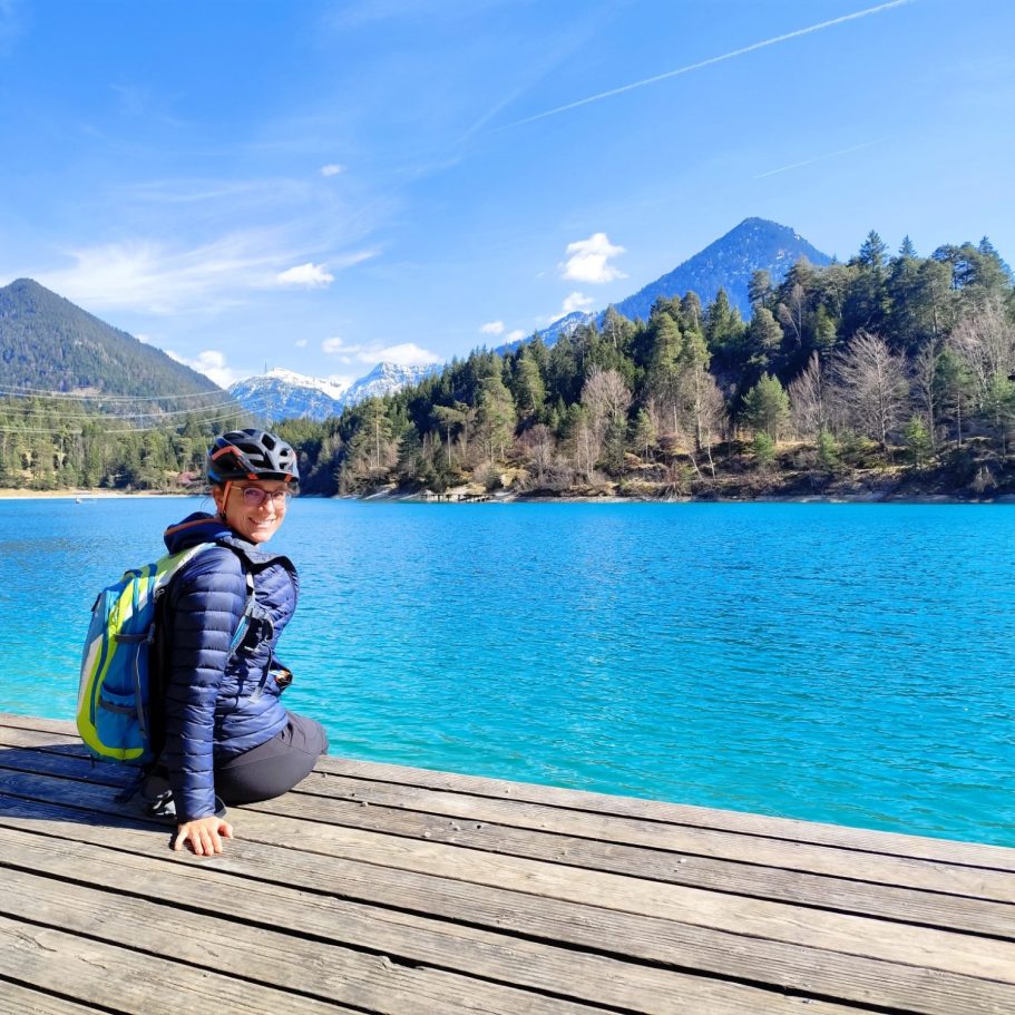 Person mit Rucksack sitzt auf einem Steg am aquablauen Wasser mit Bergen im Hintergrund.