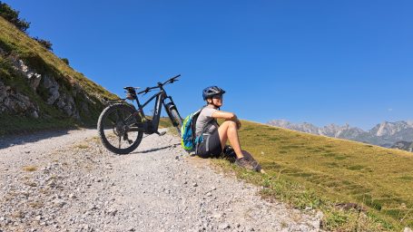 Mann sitzt neben seinem Fahrrad auf einem Schotterweg in den Bergen.