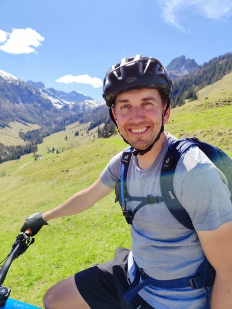 Mann mit Helm auf dem Fahrrad in einer bergigen Landschaft mit Grün und blauen Himmel.