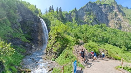 Wasserfall mit grünen Bergen und Wanderern auf einem Pfad im Vordergrund.