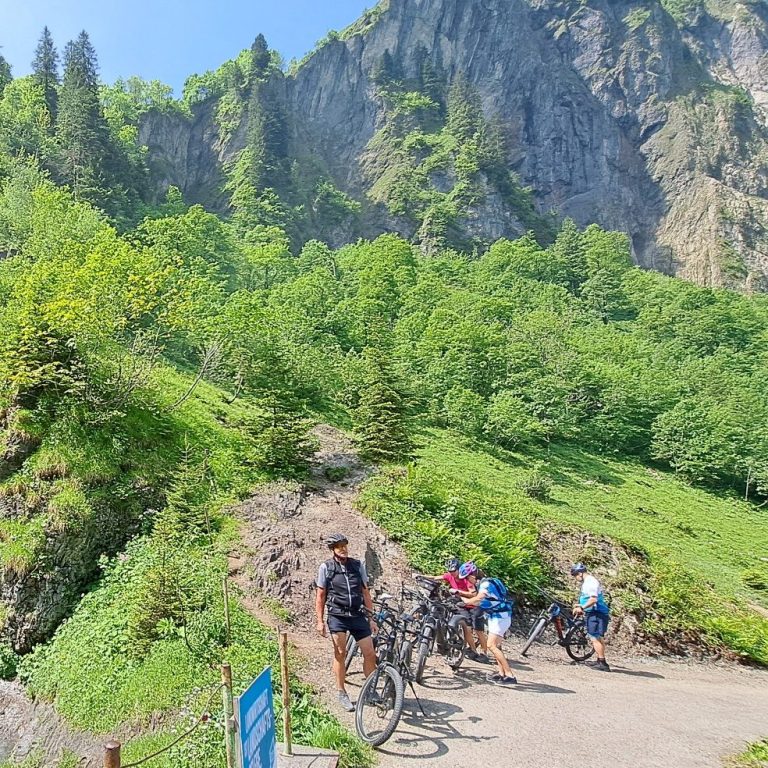 Drei Radfahrer am Sonntag auf einer grünen Bergwiese mit steilen Felsen im Hintergrund.