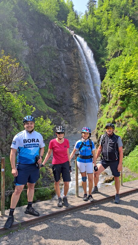 Vier Personen stehen nahe einem Wasserfall in einer grünen, bewaldeten Umgebung.