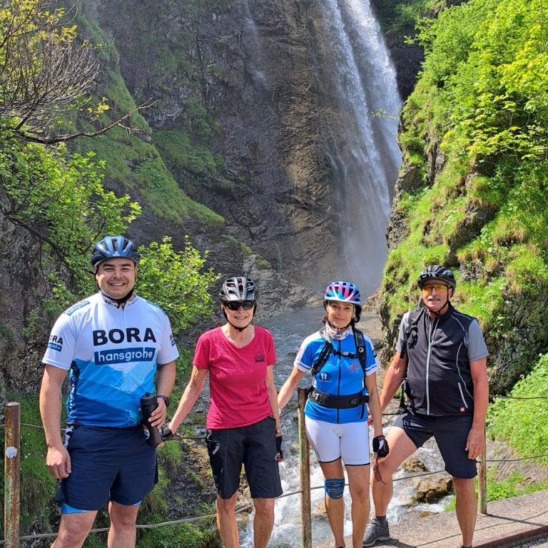 Vier Radfahrer stehen lächelnd vor einem Wasserfall in einer grünen Landschaft.