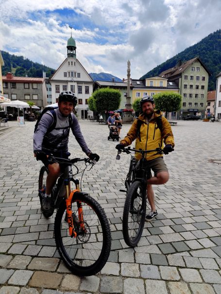Zwei Radfahrer in einer Stadt mit historischen Gebäuden und Bergen im Hintergrund.