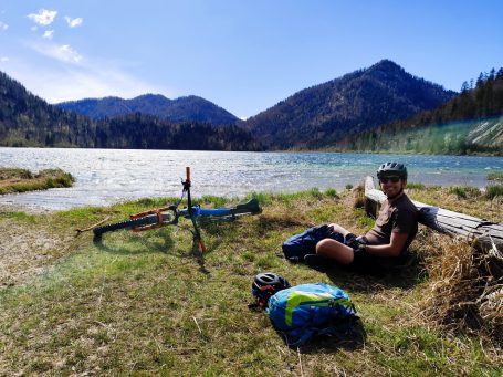 Junger Mann sitzt am Ufer eines Sees mit einem Fahrrad, umgeben von Bergen.