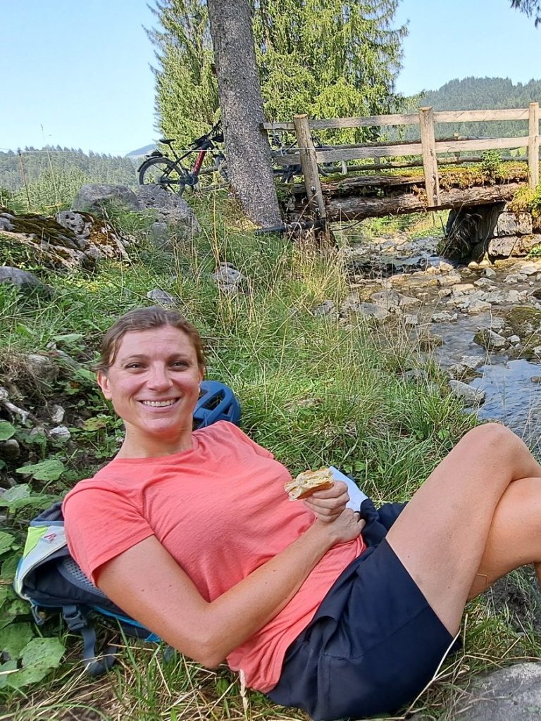 Frau sitzt entspannt am Fluss, hält ein Sandwich und lächelt in die Kamera.