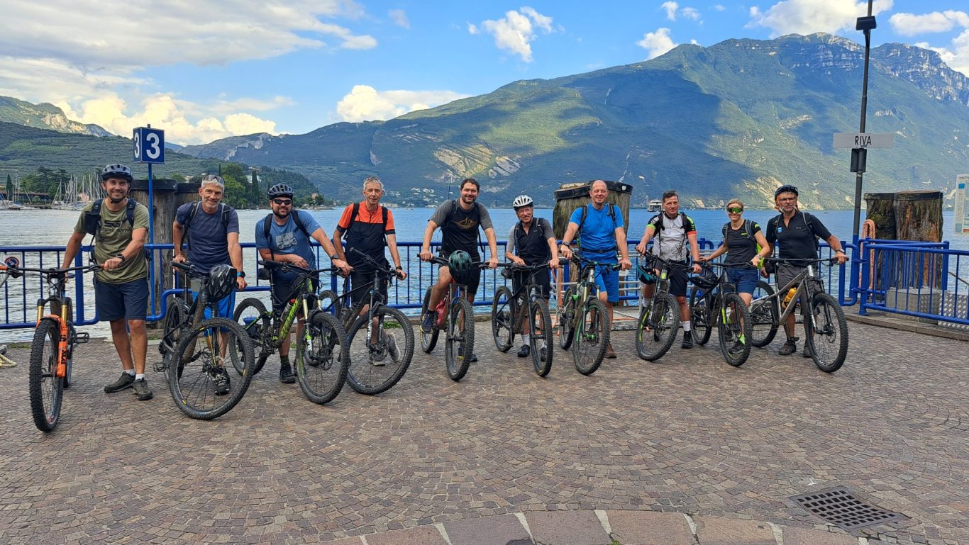 Finisher am Gardasee Gruppe von Radfahrern steht an einem Ufer mit Bergen im Hintergrund.