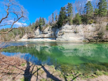 Grüner Fluss mit umliegenden Felsen und Bäumen unter blauem Himmel.